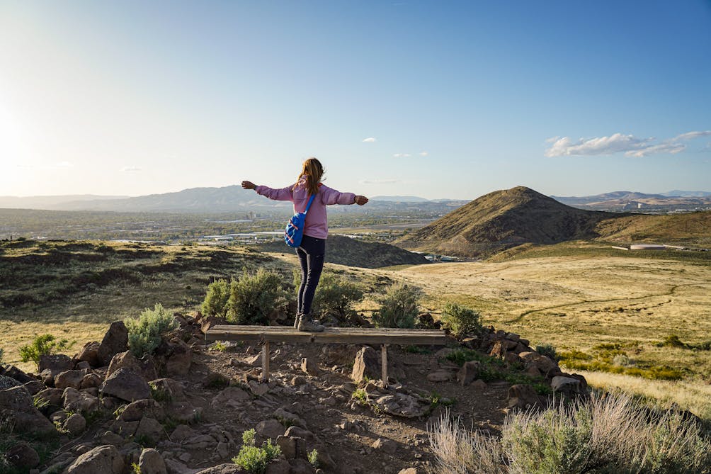 A woman is standing with her arms spread out as she takes in the view of mountains and downtown Reno at Huffaker Hills Open Sapce 