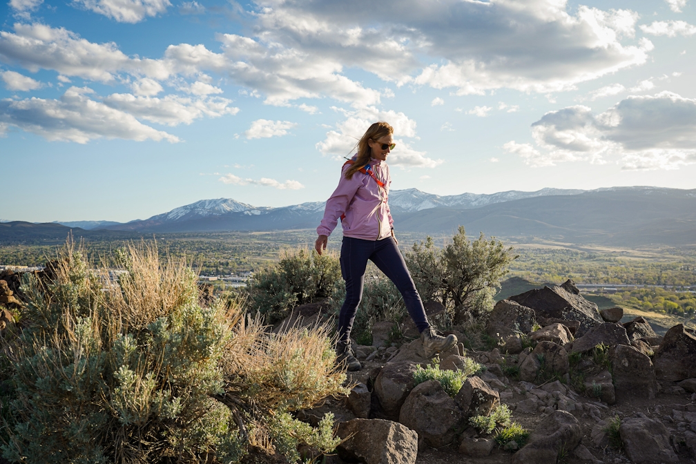 A woman in a pink windbreaker jacket is standing atop a rocky outcropping Huffaker Hills Open Space in Reno