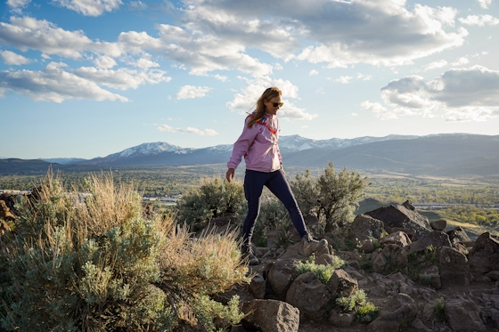 A woman in a pink windbreaker jacket is standing atop a rocky outcropping Huffaker Hills Open Space in Reno
