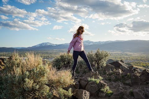 A woman in a pink windbreaker jacket is standing atop a rocky outcropping Huffaker Hills Open Space in Reno 