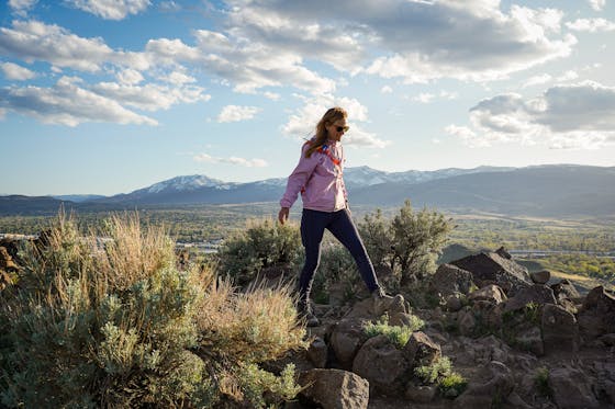 A woman in a pink windbreaker jacket is standing atop a rocky outcropping Huffaker Hills Open Space in Reno 