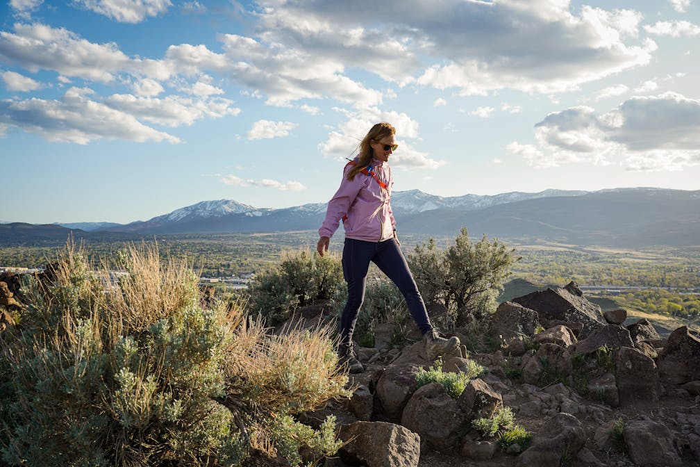 A woman in a pink windbreaker jacket is standing atop a rocky outcropping Huffaker Hills Open Space in Reno 