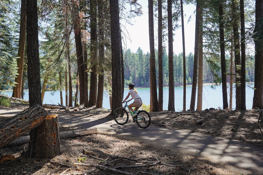 A woman is riding her bike past trees and Lake Almanor on the Lake Almanor Recreation Trail near Chester, California