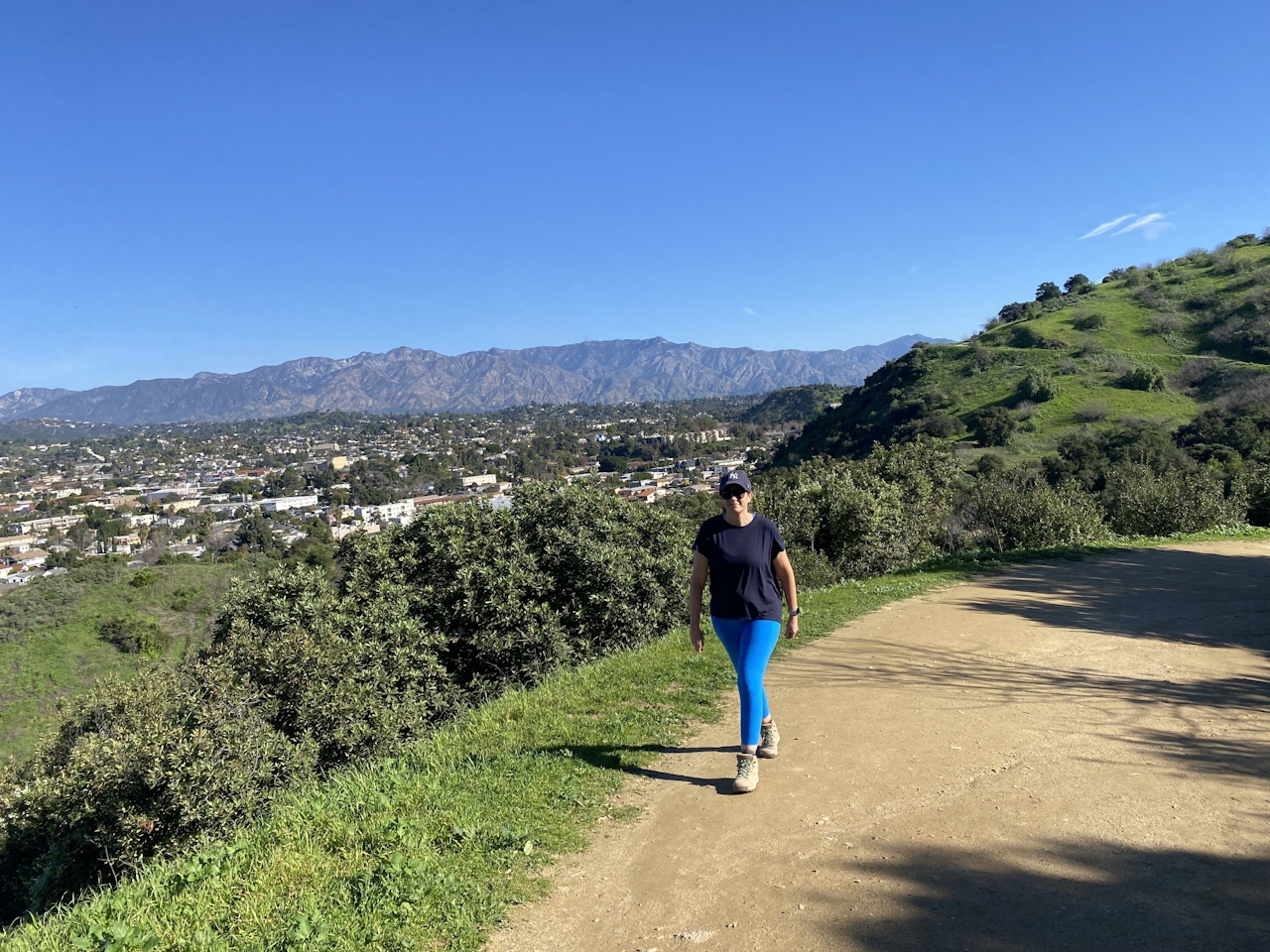 A woman hikes a wide dirt trail with green hills beside her and mountains in the background as well as city views of Los Angeles. This is at Ernest B Debs Regional Park in Los Angeles