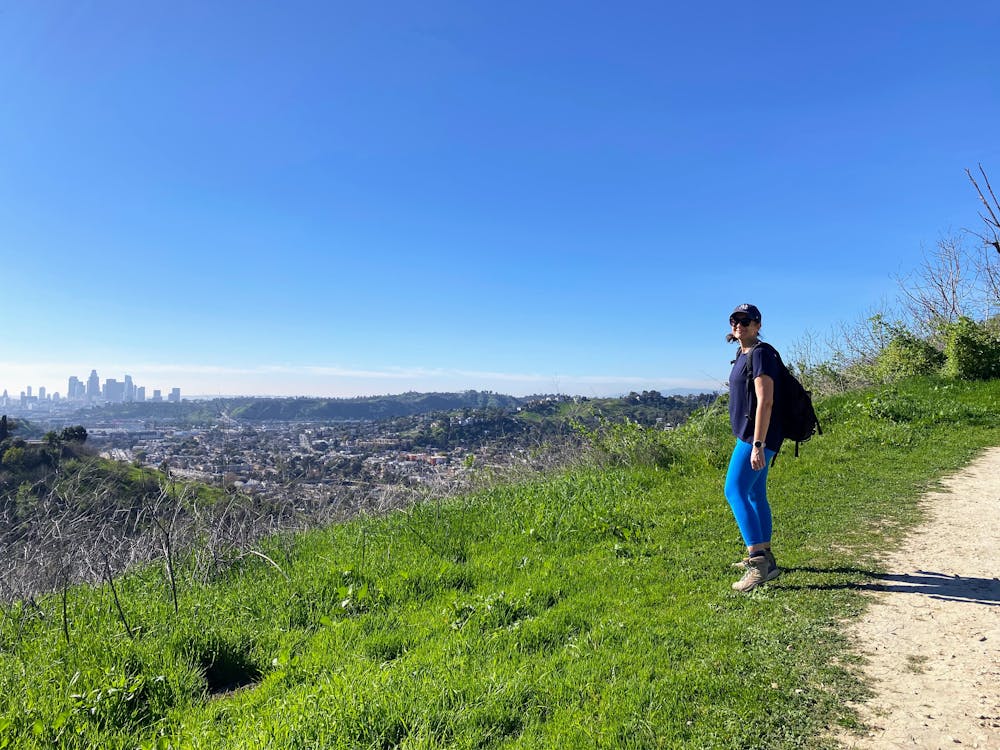 A woman stands on green grass overlooking the skyline of downtown Los Angeles from Ernest E Debs Regional Park