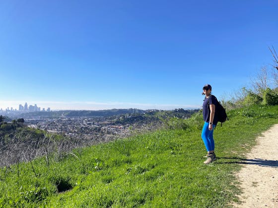 A woman stands on green grass overlooking the skyline of downtown Los Angeles from Ernest E Debs Regional Park