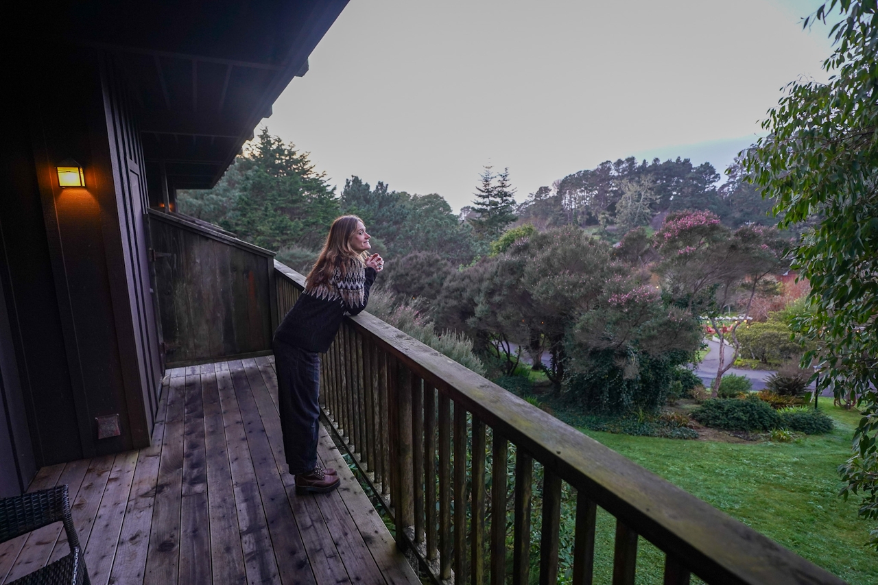 A woman stands on a hotel room balcony overlooking the lush green grounds at Stanford Inn at the Sea in Mendocino