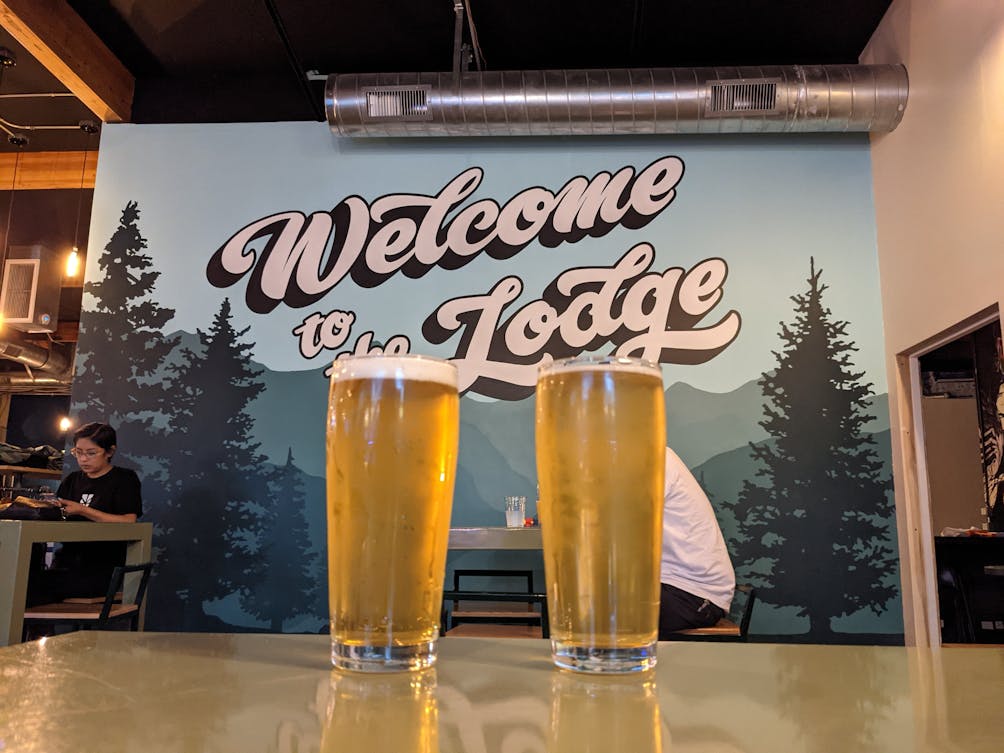Two beers in glasses set in front of a Welcome to the Lodge sign at Arrow Lodge Brewing 