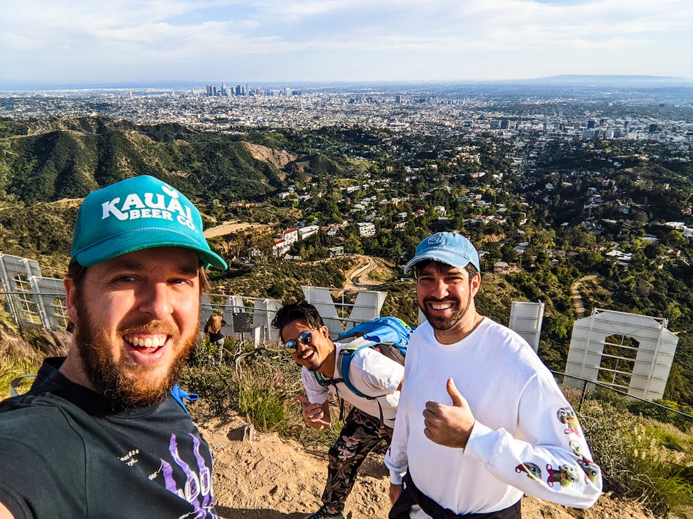 Three guys taking a selife and smiling and giving the thumbs up from behind the famous Hollywood Sign 
