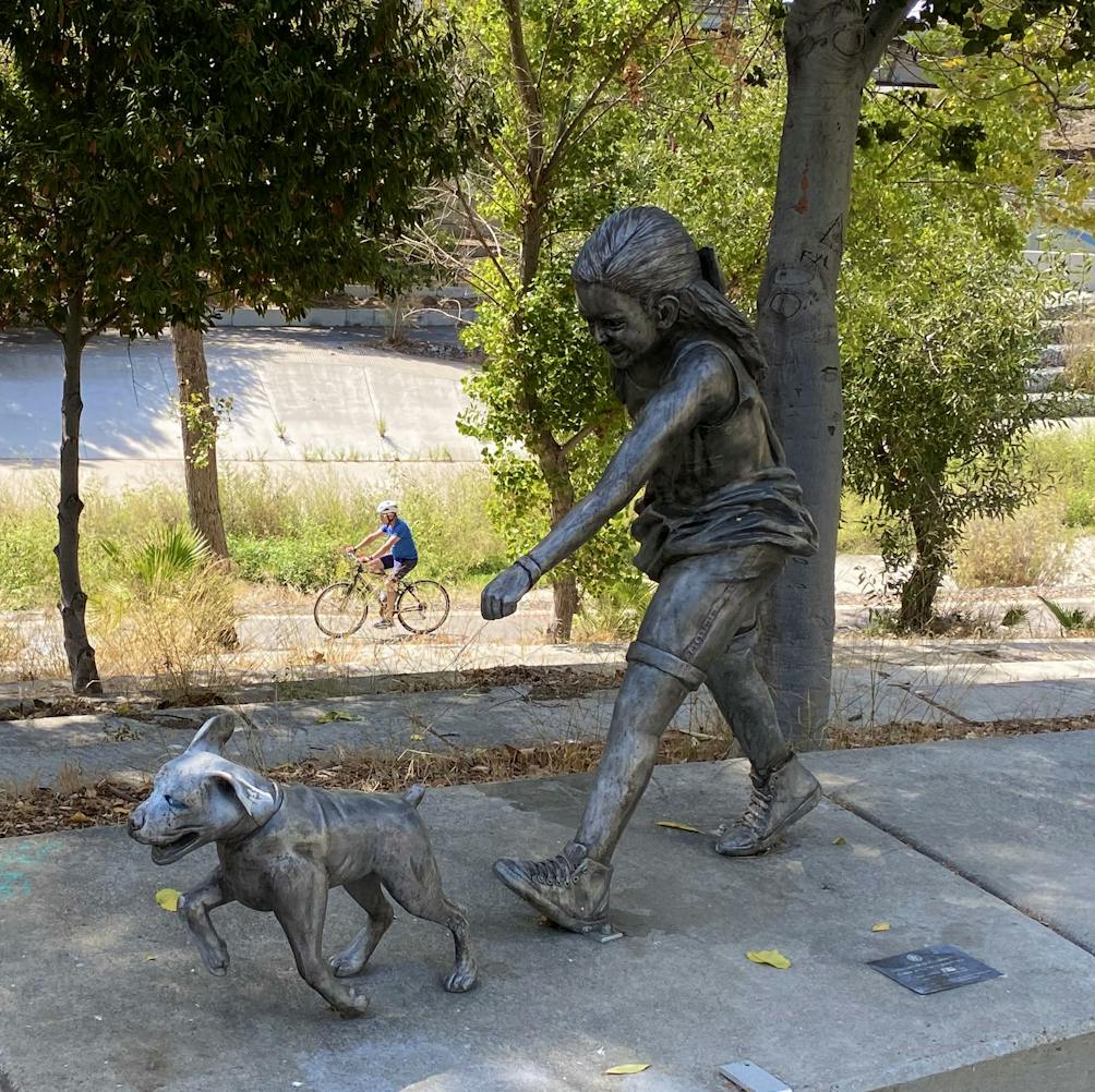 sculpture of a young girl walking her dog on a sidewalk with bike riders on the bike path below in San Jose at Guadalupe River Trail 