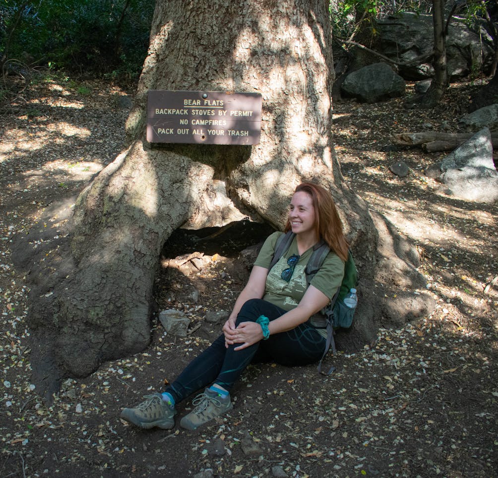 Woman taking a hiking break and sitting by a tree in the shade and the tree has a sign marking it as Bear Flats Backcountry Campground on Mount Baldy in Los Angeles County 