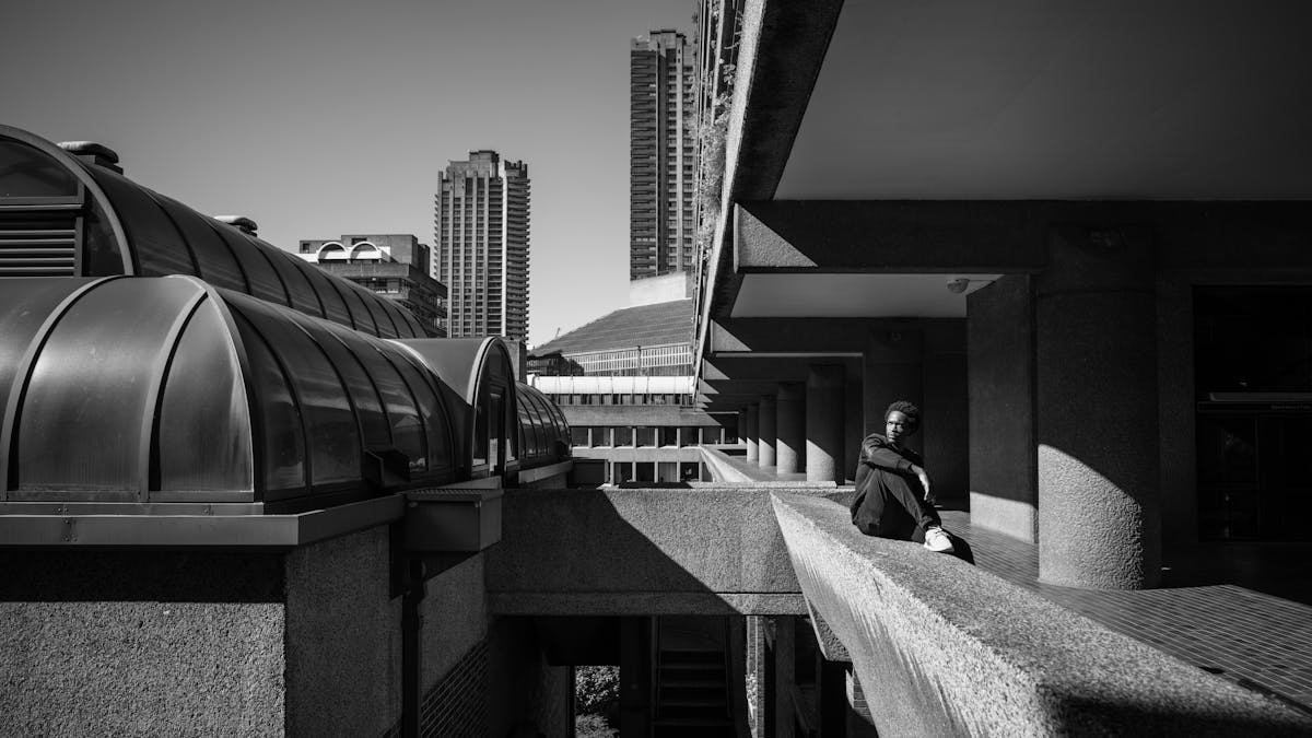 Photograph in black and white of a man sitting on a walkway balcony looking out at the Barbican Centre, London.
