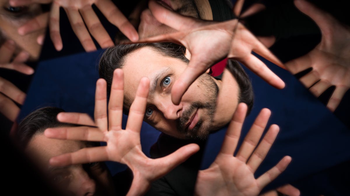 Photograph through a kaleidescope of the head of a man. His hands are held up in front of his face, with fingers outstretched, framing has eye. The background is made up of blue and red fabric.