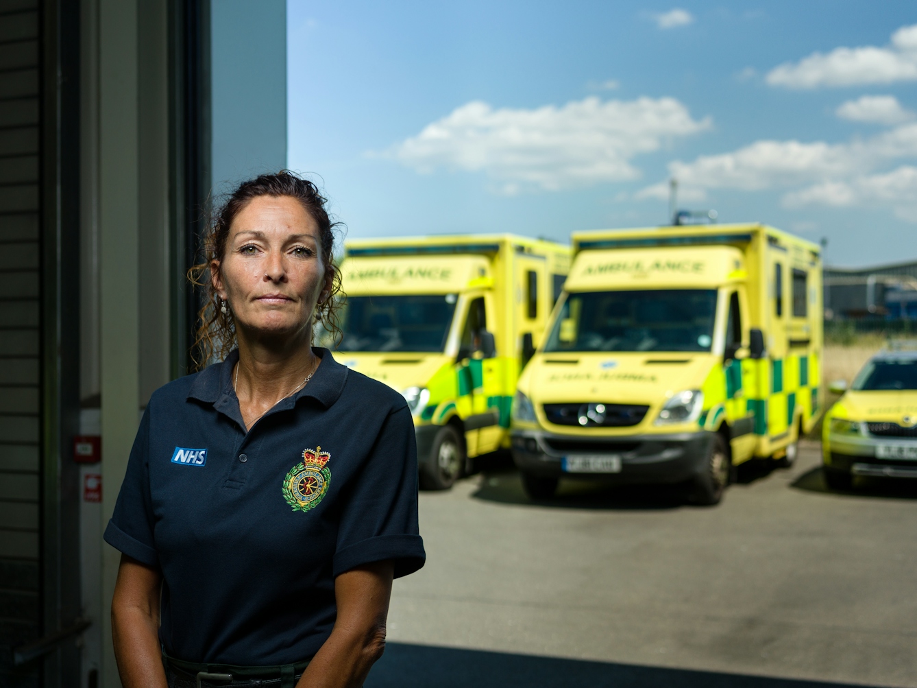 Photographic portrait of Mandy English, paramedic, at Hull East Ambulance Station, with ambulances in the background.