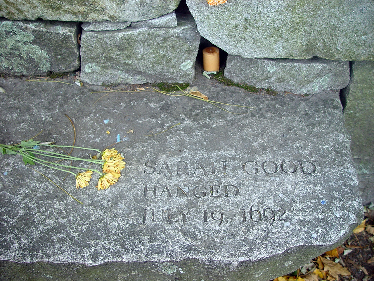 Colour photograph of a grey stone with wilted yellow flowers upon it and an inscription that reads "Sarah Good hanged July 19, 1692".