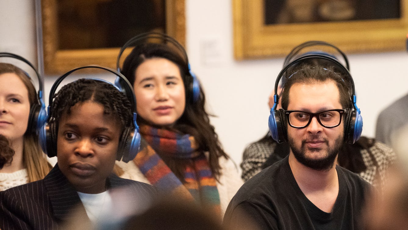A photograph of a group of people at an event in the Reading Room in Wellcome Collection. They are all wearing headphones. Behind them are paintings on the wall. 