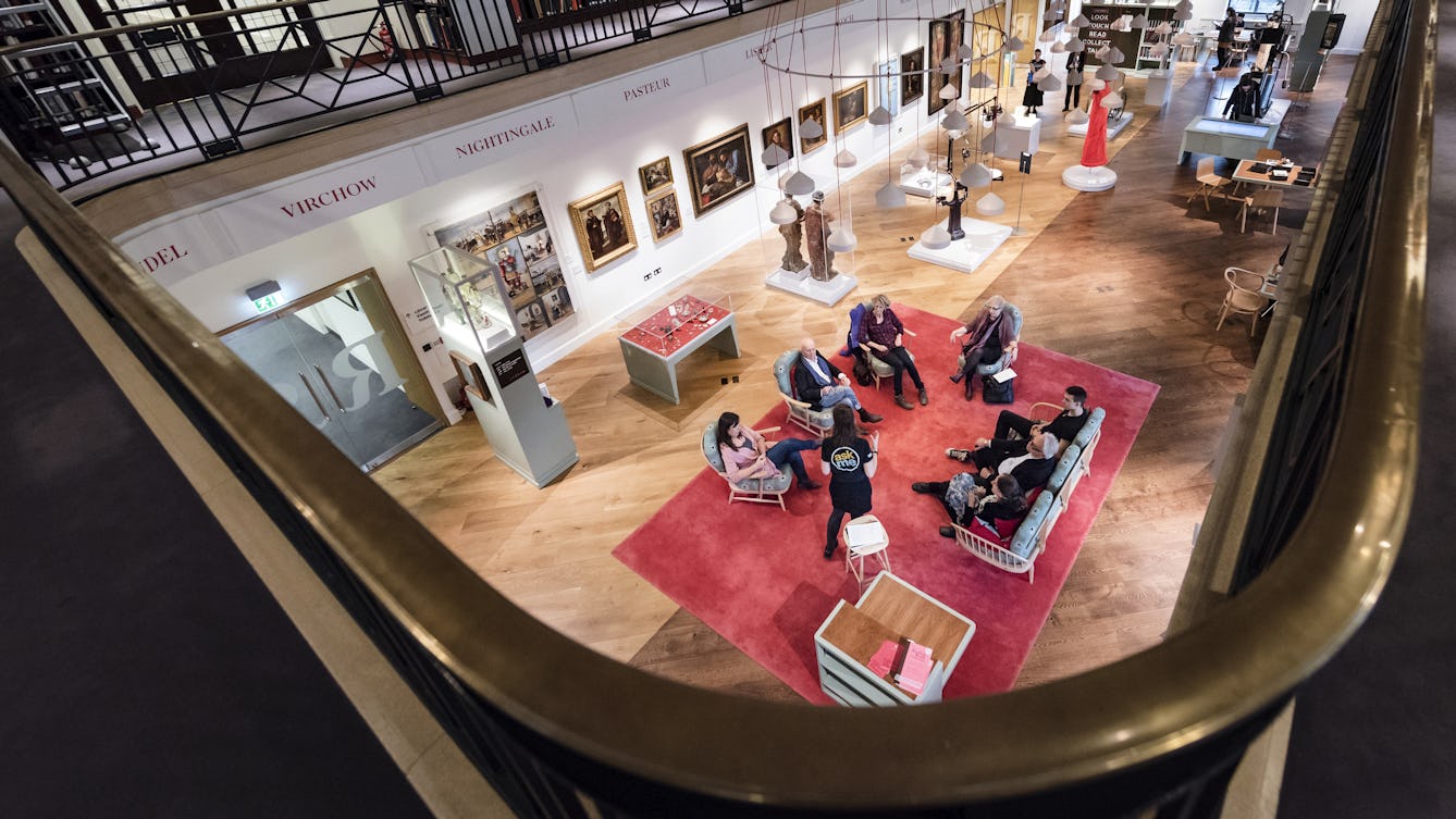 Colour photograph taken from the Wellcome Reading Room balcony looking down into the Reading Room, where a group of people are sitting in a circle listening to a Visitor Experience Assistant who is standing in front of them wearing a black t-shirt.