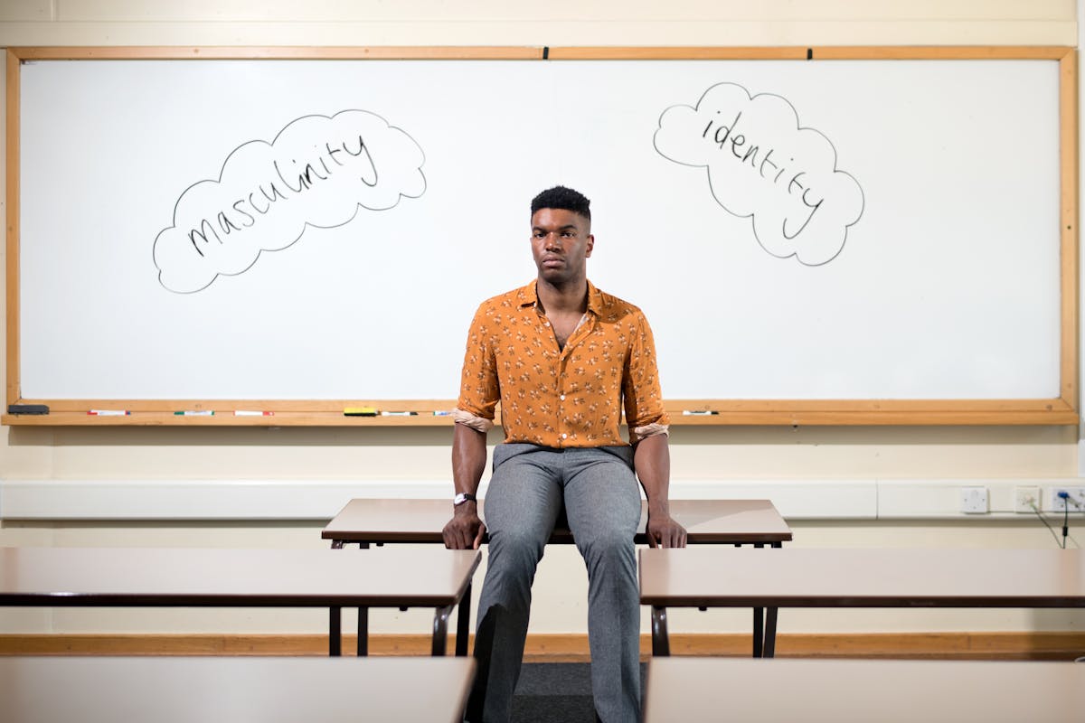 Photograph of a man in an orange shirt and grey trousers sat on a desk in a classroom. behind him on the white board is written the words, 'masculinity' and 'identity'.
