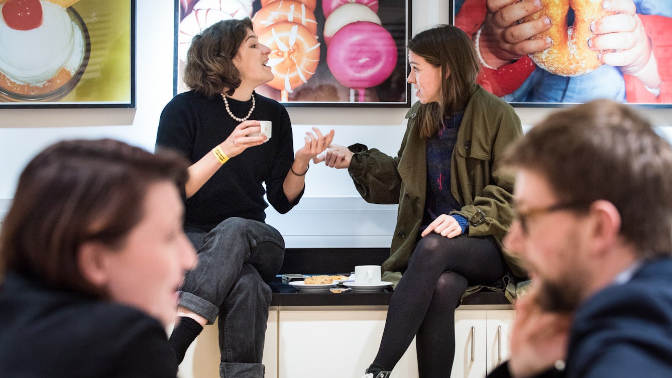 Photograph of two young women in discussion, seated in front of colourful photographs of food.