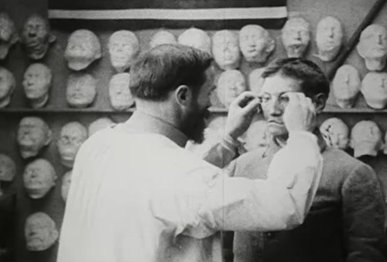 A man in a white coat adjusts the spectacles on another man's face.