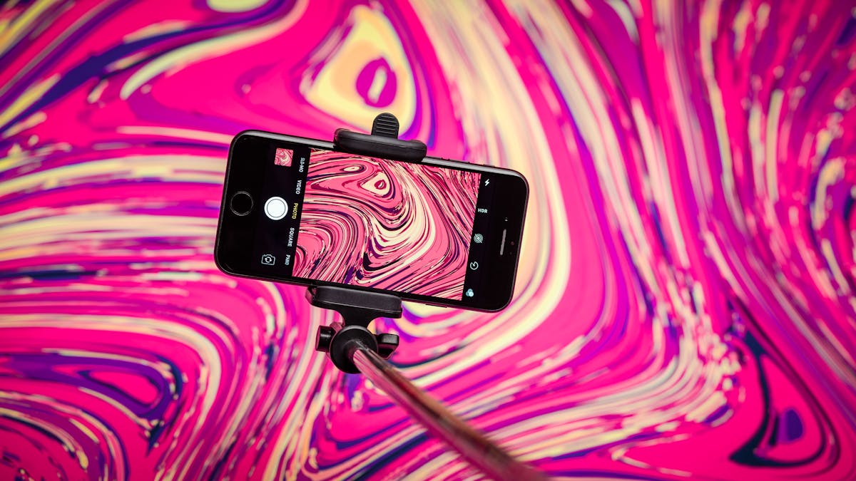 Photograph of a smart phone held in a selfie-stick, in front of a colourful swirly background. The background pattern is repeated on the phone screen. 