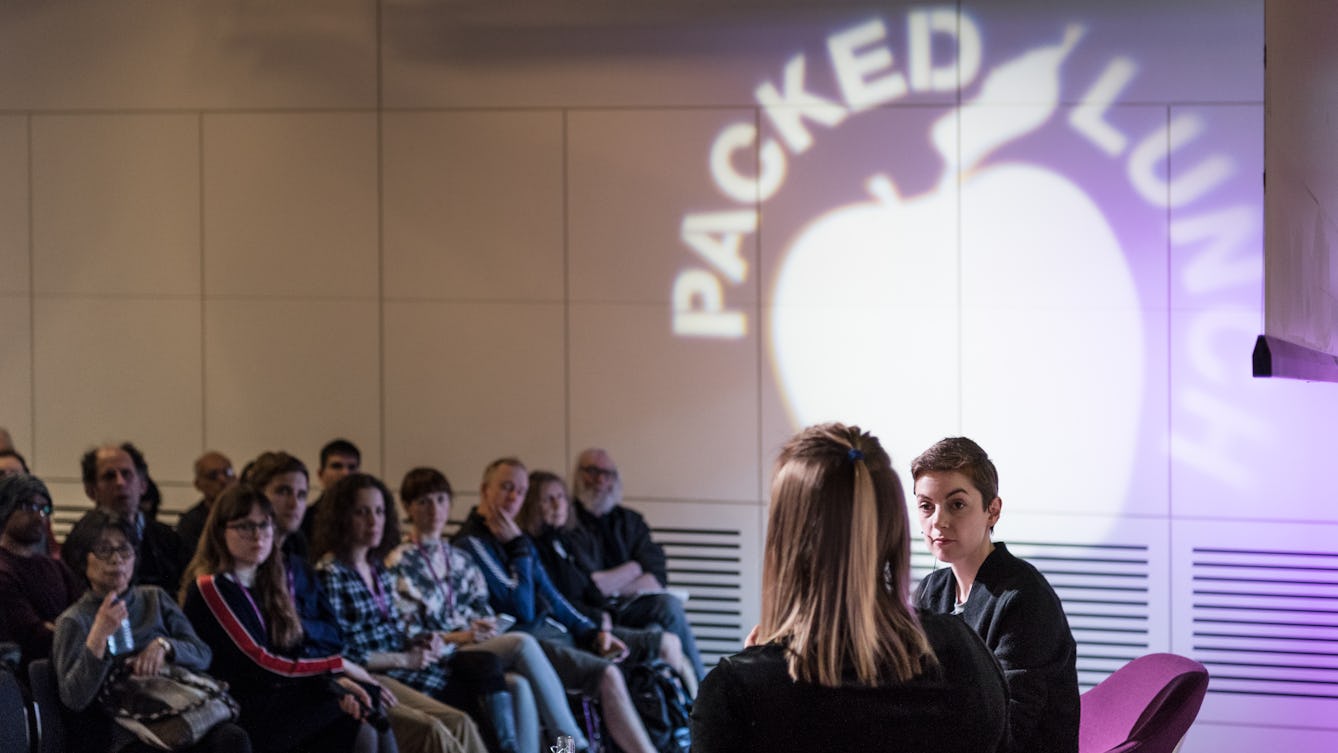 Photograph of two women having a conversation on a small stage. In the background are the audience. Projected onto the far wall is the silhouette of an apple with the words Packed Lunch circling the top of the apple. 