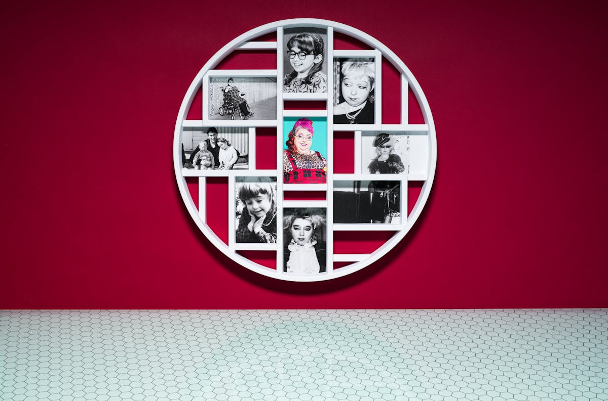 Photograph of a multiframe photo frame containing nine photographs, one in colour and eight in black and white. The frame is hung on a crimson wall above a glass patterned tabletop.