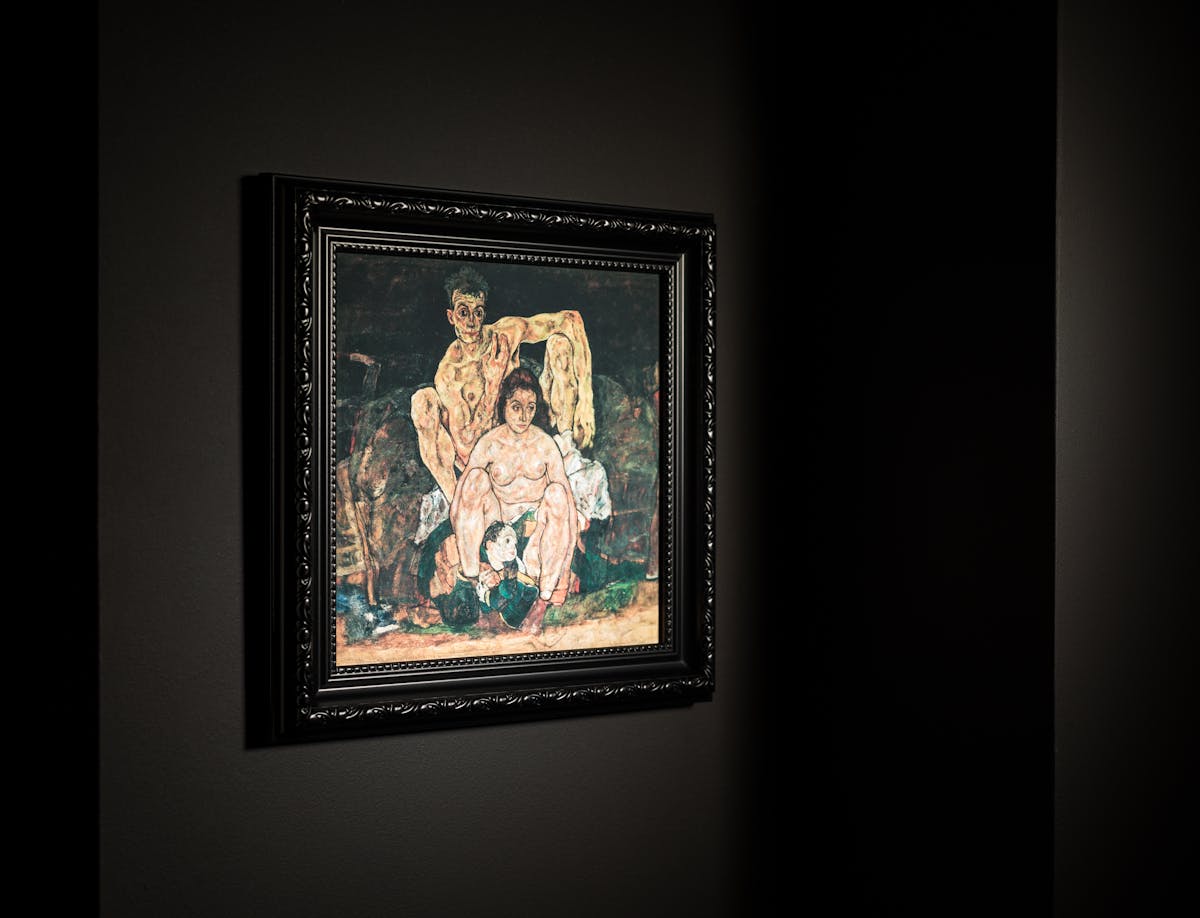 Photograph black ornate picture frame hung on a black wall. Inside the frame is a print of an oil painting by Egon Schiele titled, 'The Family'. It depicts  Schiele himself at the far back, his sinewy nude body hunched behind his wife, Edith, who looks off to the side, while a child is curled between her feet.