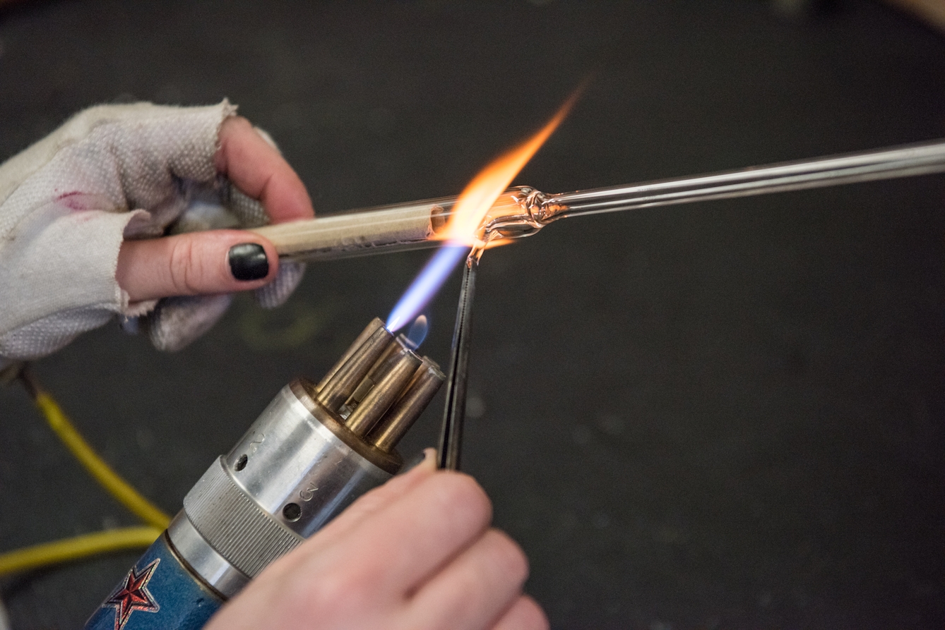A hand in protective glove with thumb and forefinger cut away holds a glass rod above the flame of a blowtorch.