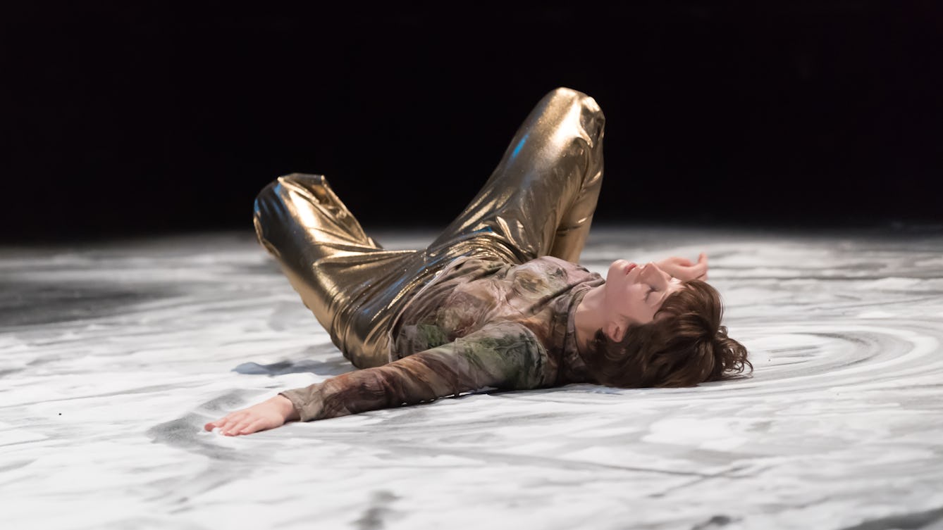 Photograph of a dancer lying on a salt-covered floor. They are wearing gold trouser and a patterned top with muted colours. Their eyes are shut and the dance has made patterns in the salt on the floor. 
