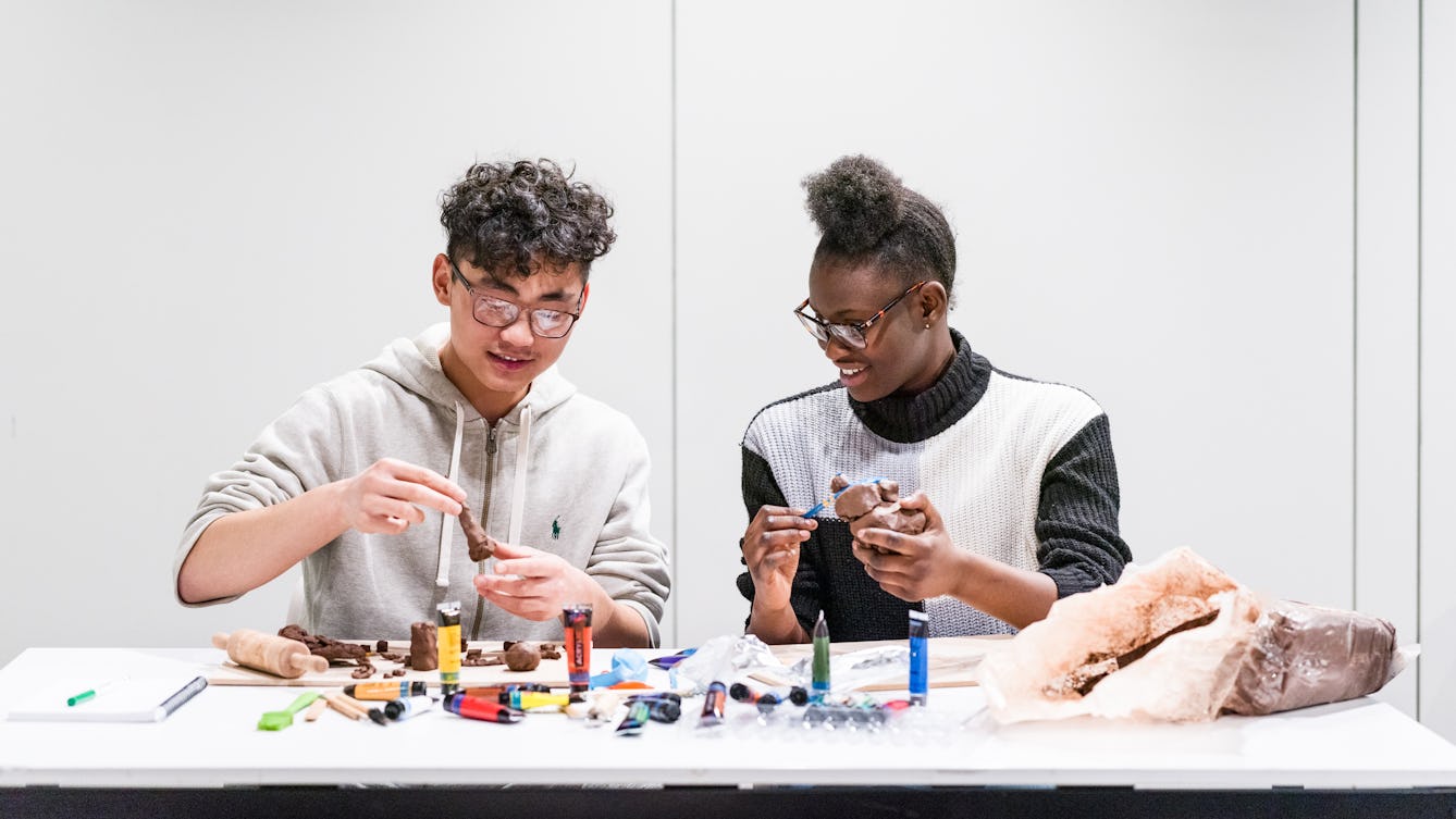 Photograph of a young man and woman sitting side by side, taking part in a table based workshop involving paints, clay and small sculptures.