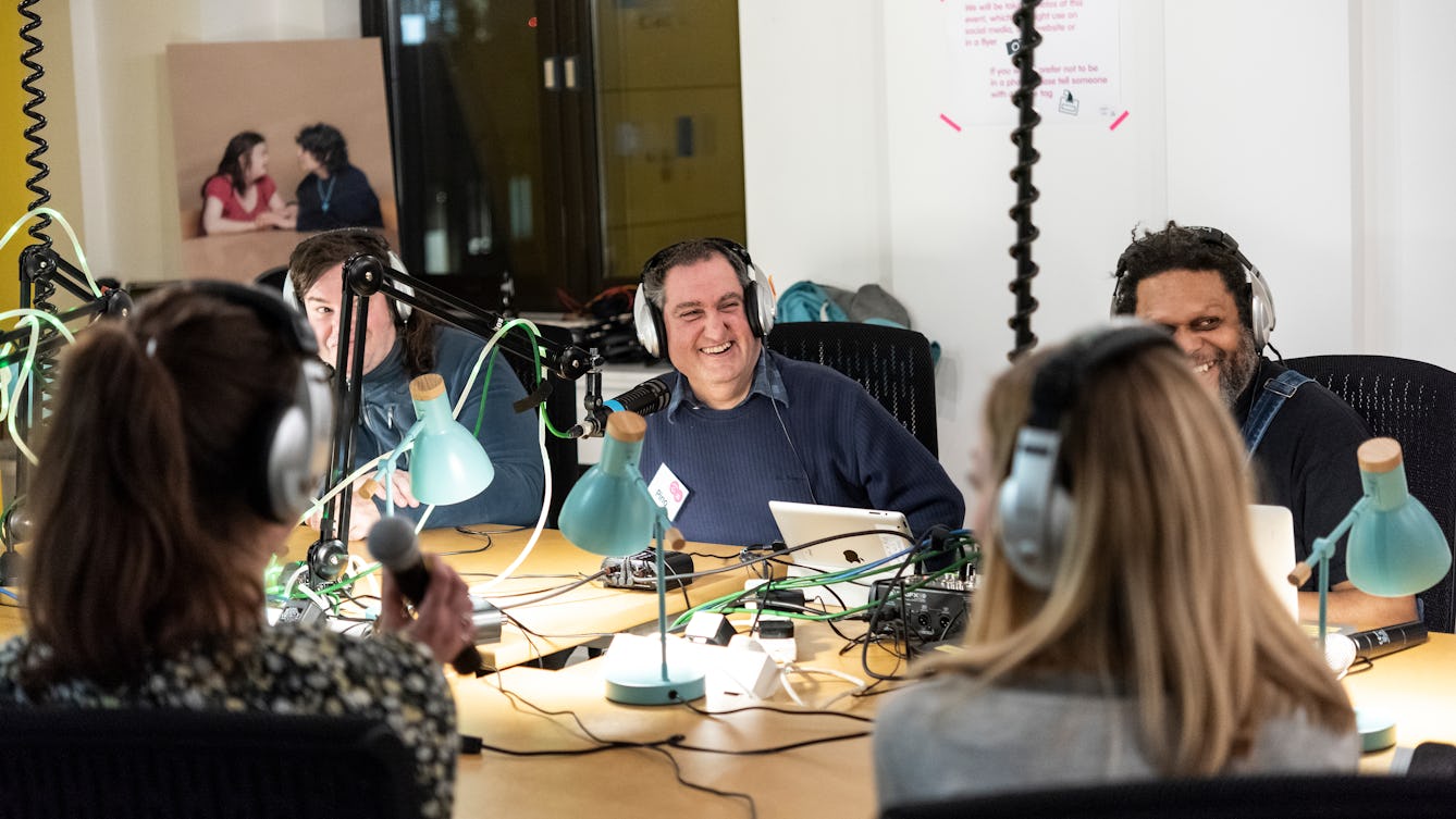 A group of people smiling and talking at a live radio session sitting around a table with microphones on the table in front of them and headphones on. 