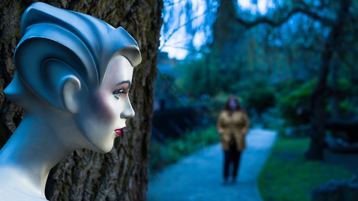 Photograph of the profile of a mannequin's head in front of a tree trunk. In the background an out-of-focus young woman approaches.