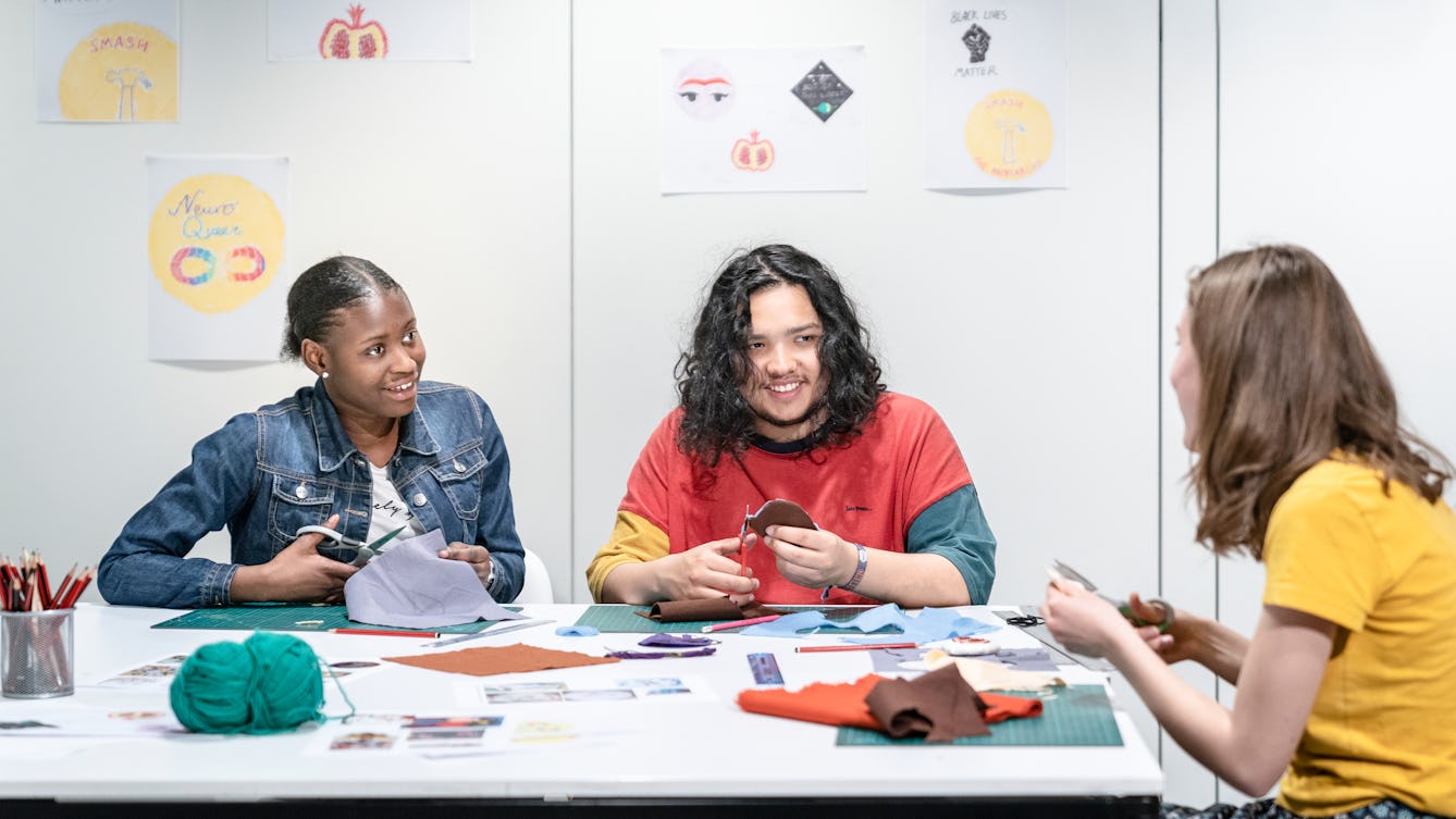 Photograph of three young participants sat at a table. The participants are cutting out felt with scissors and talking to eachother. On the wall behind them are illustrations. 