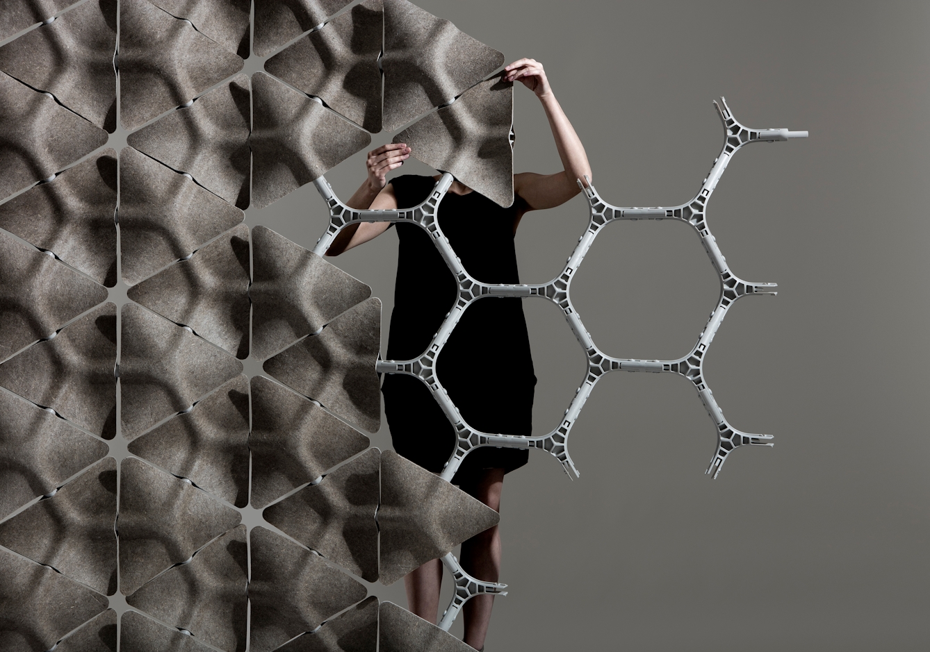 Colour photograph of a person behind a hexagonal grey plastic frame, who is in the process of affixing an grey rough-textured triangle to form a wall with other tessellated tiles on the frame.