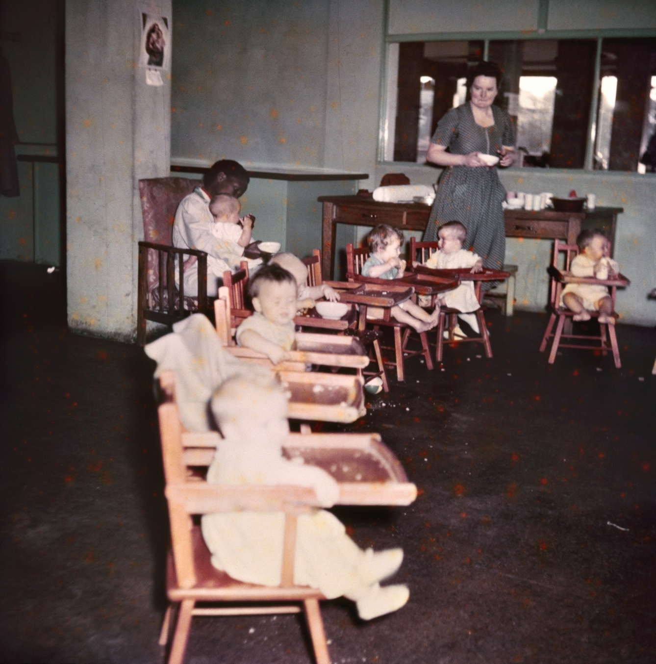 Photograph from the mid-20th Century Peckham Pioneer Centre, showing a long row of eight high-chairs, six of which are occupied by infants who are eating or drinking. Behind them, a woman sits in a chair with a child on her lap and a bowl in her hand, and another woman approaches from behind holding another bowl.