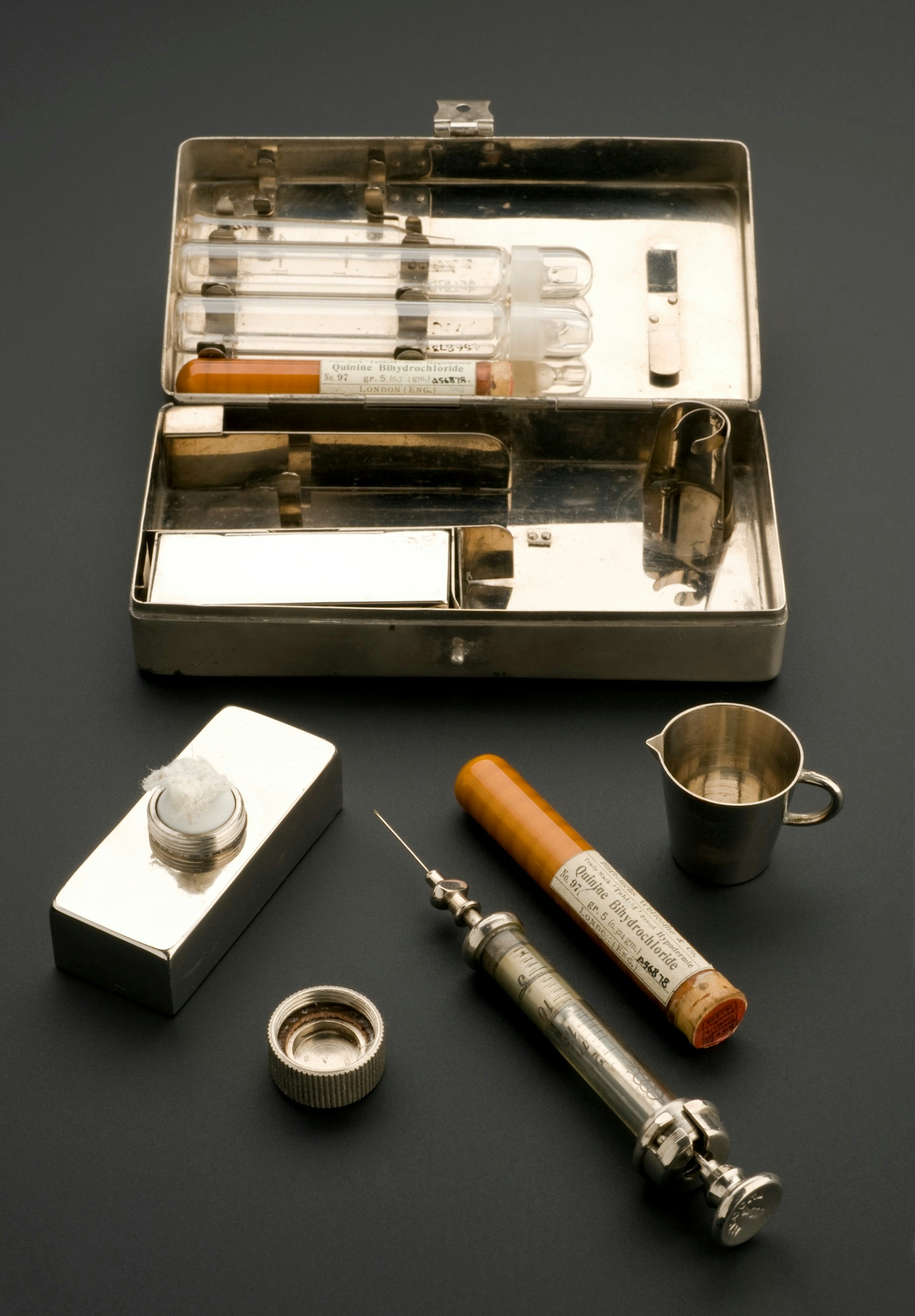 Rectangular metal box containing empty phials. in front of the case, its other contents are laid out: a bottle-shaped container holding cotton wool, a metal syringe with glass body, a tube of quinine bihydrochloride and a small metal jug.