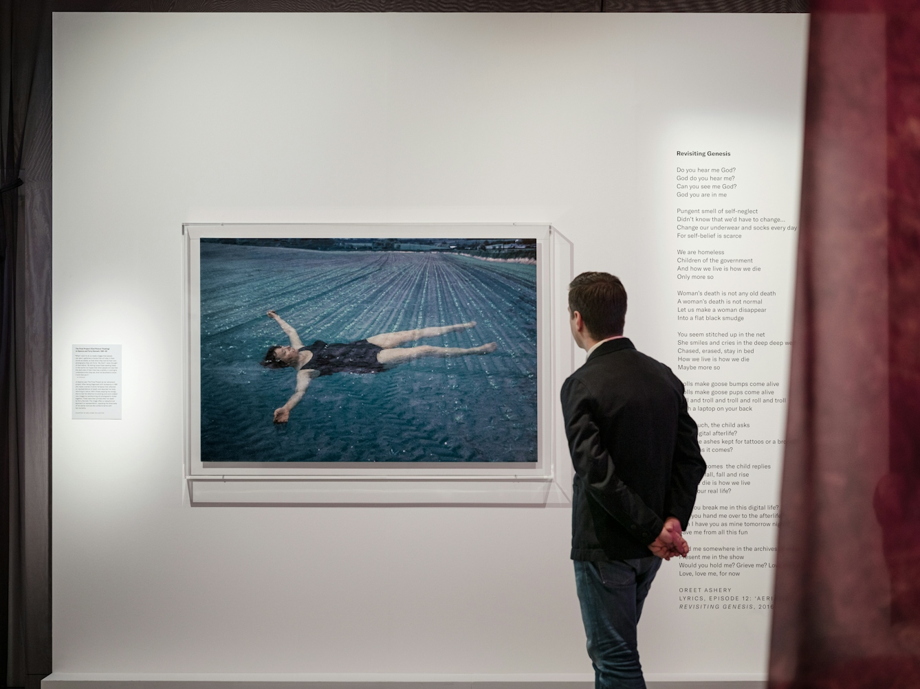 Photograph of a man in a dark jacket and jeans viewing a photograph of a woman floating belly-upwards in a blue pool. To the right of the man there is a poem called Revisiting Genesis