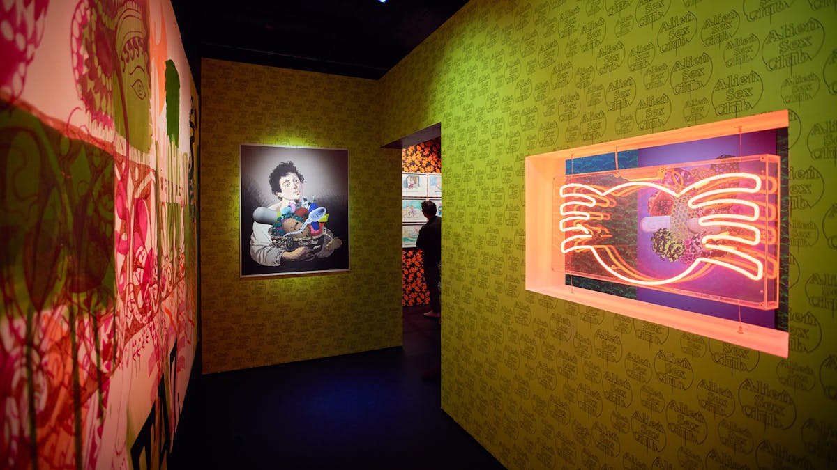 Photograph of a visitor in the distance looking at framed artworks on the wall. In the foreground is colourful patterned wallpaper, a work of art and a red neon exhibit.