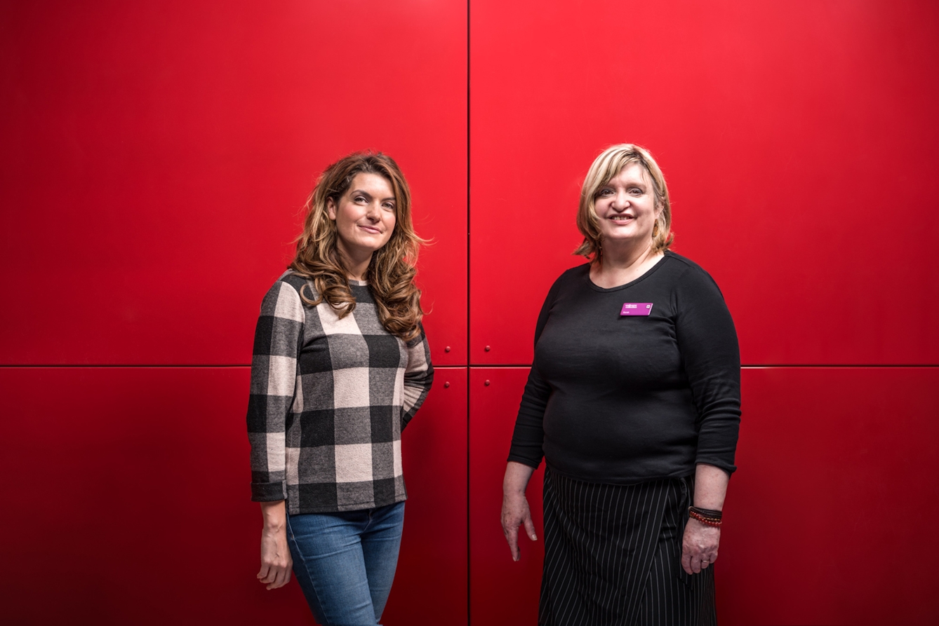 A portrait of two women standing against a bright red background.