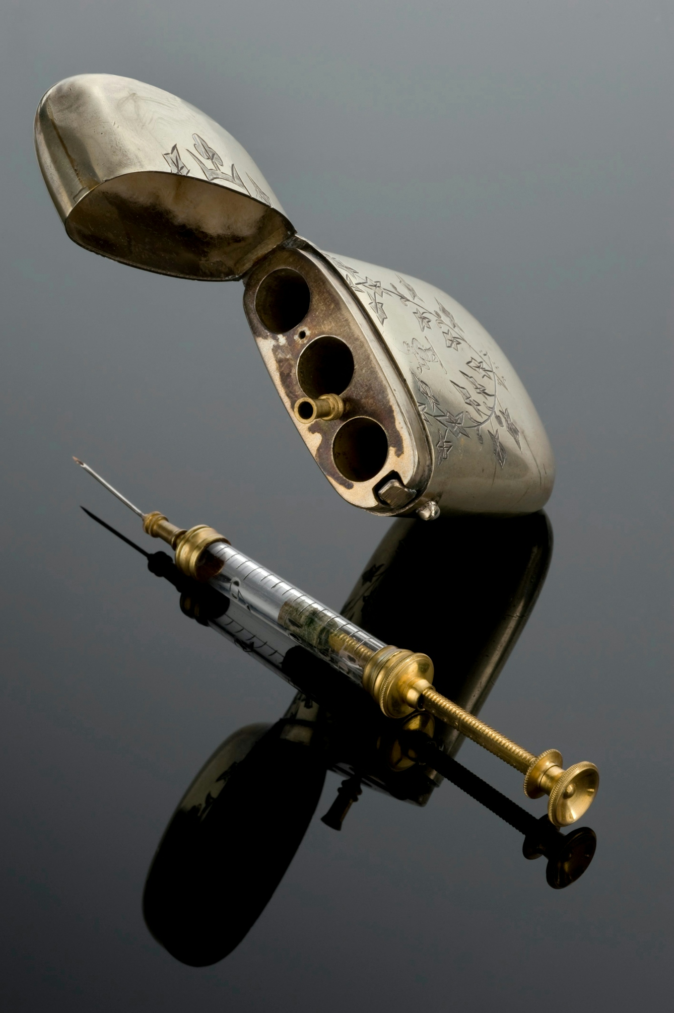 Gold-coloured syringe with a glass tube with measure-lines on, and an engraved rounded metal case similar to a cigarette-case.