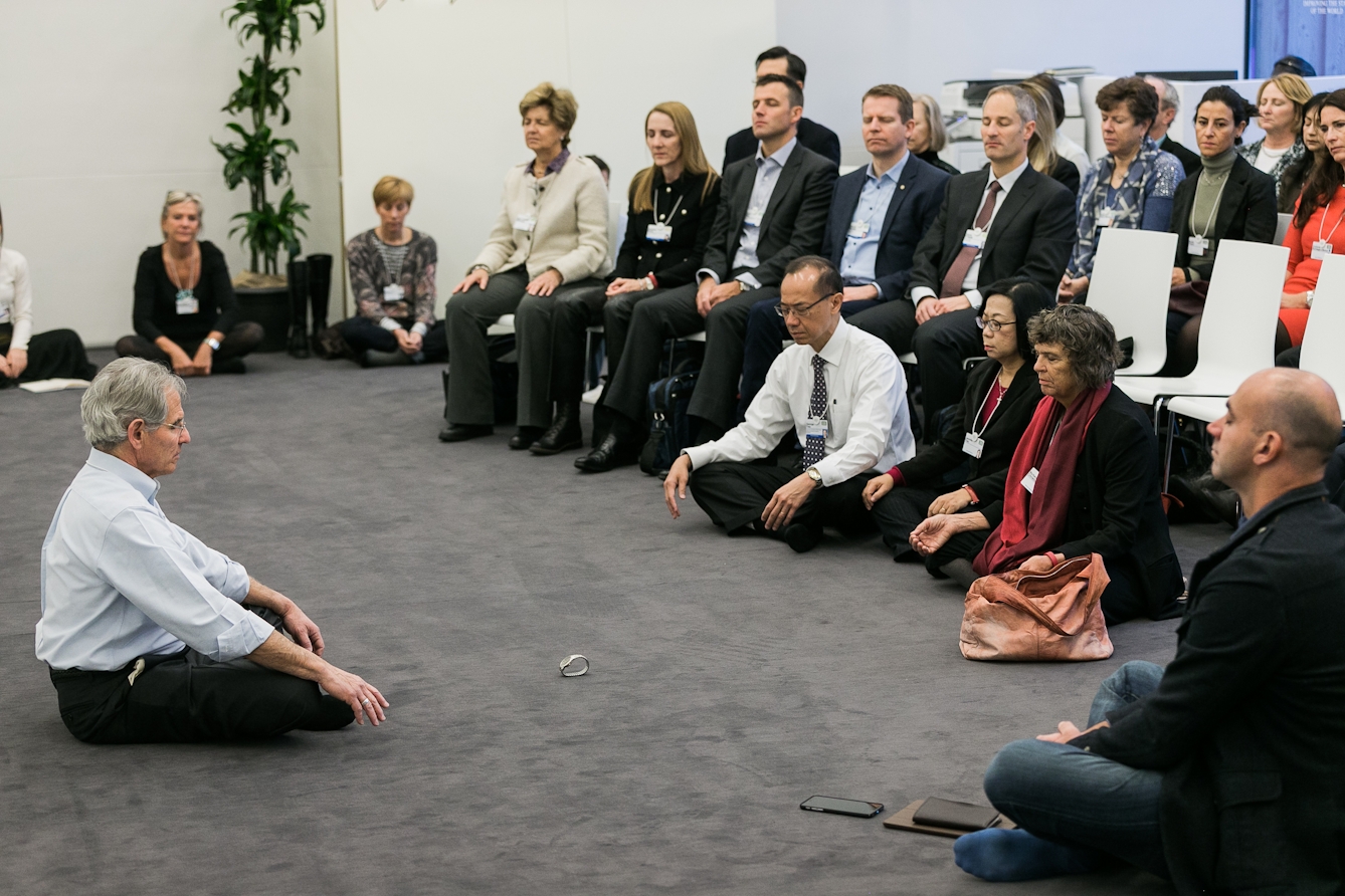 A grey haired man wearing glasses, a button-down shirt and suit trousers sits cross-legged in front of a group of people in business attire. All have their eyes closed in mindfulness meditation.