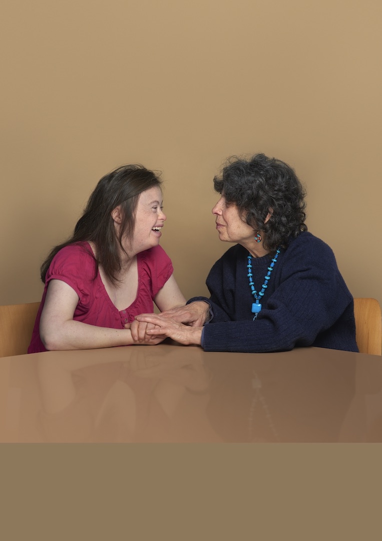 Colour photograph portrait of two women sitting side-by-side, Chloe and Zelda McCullum. They sit at a table looking affectionately at each other and Zelda covers Chloe's hands with her own.