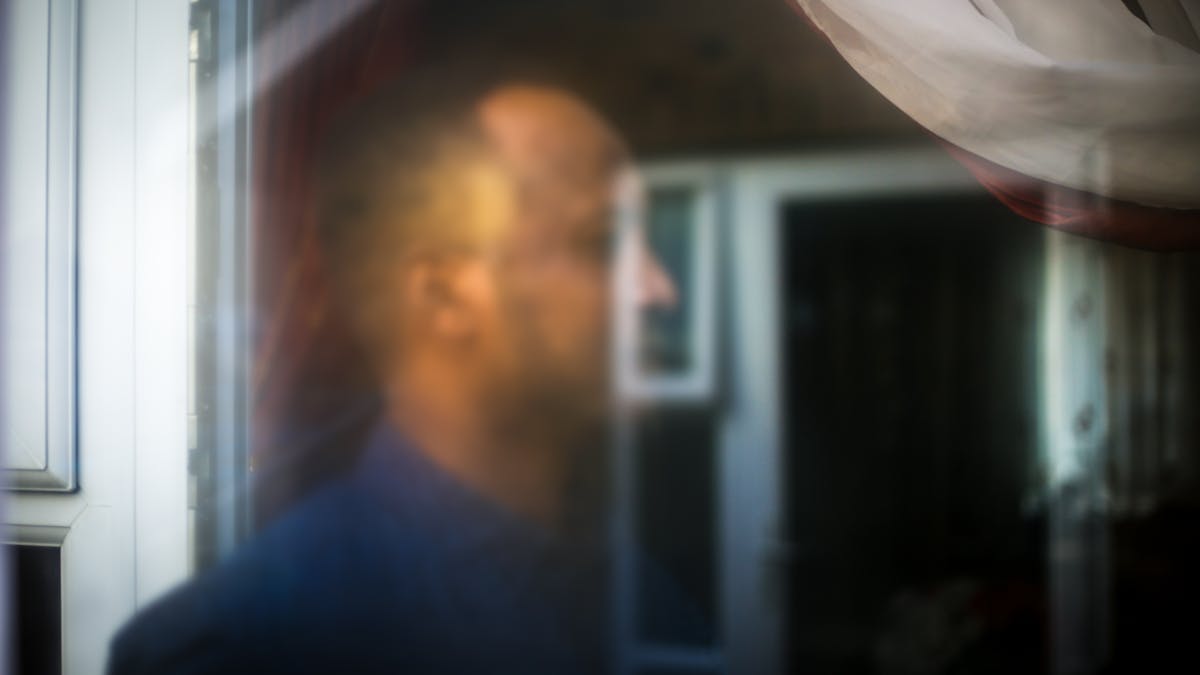 Photograph of a man's head seen through a glass window. His head is out of focus and merges with the reflection of another scene.