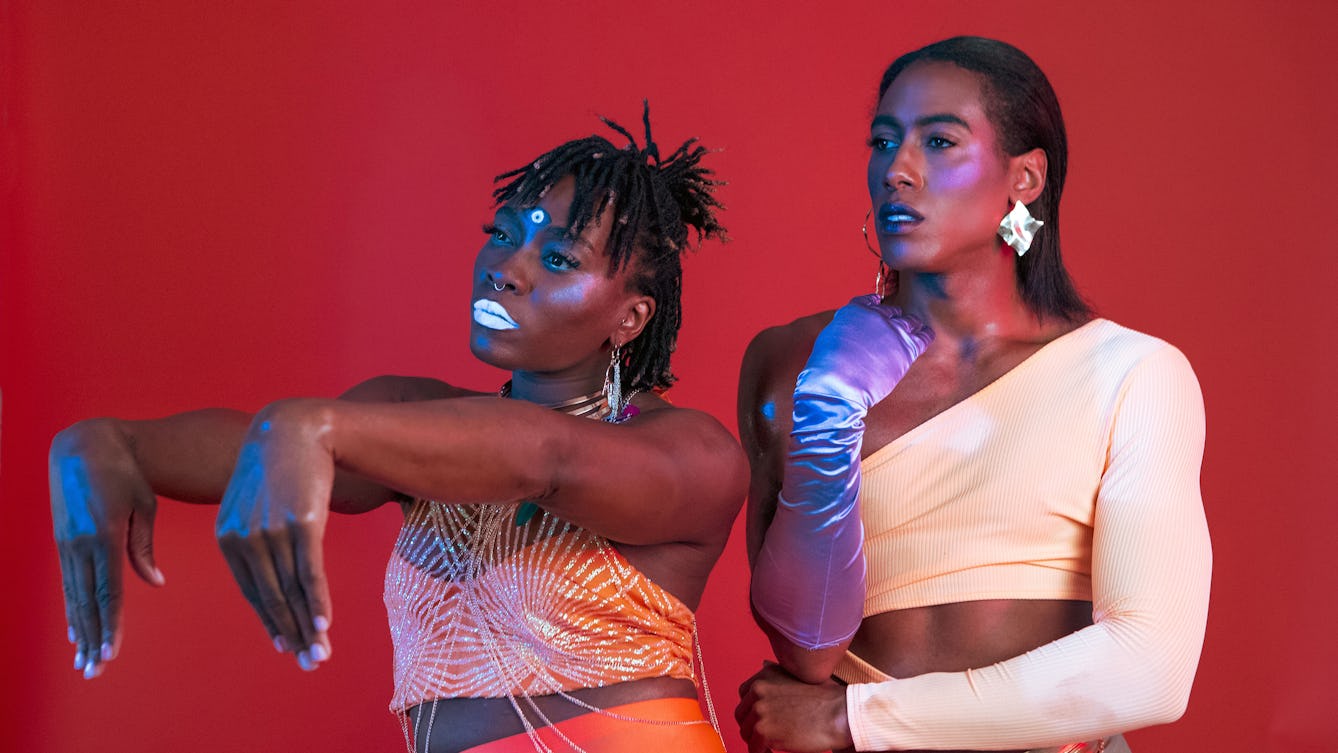 Two artist performers posing in luminous make-up and clothes against a red background.