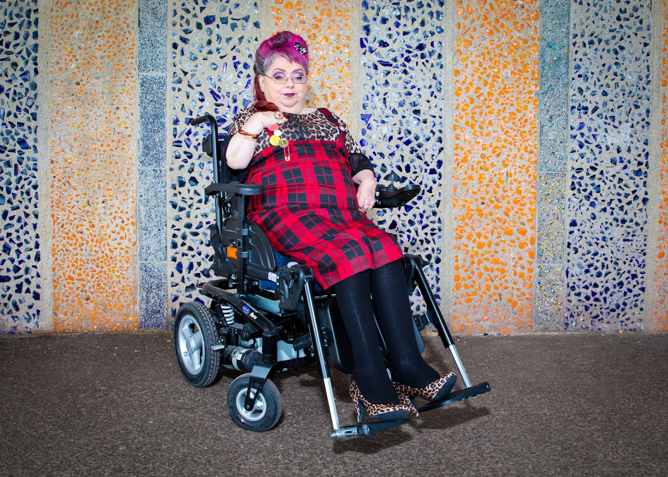 Photograph of a woman wearing a red tartan dress seated in a wheelchair, against a coloured mosaic patterned background.