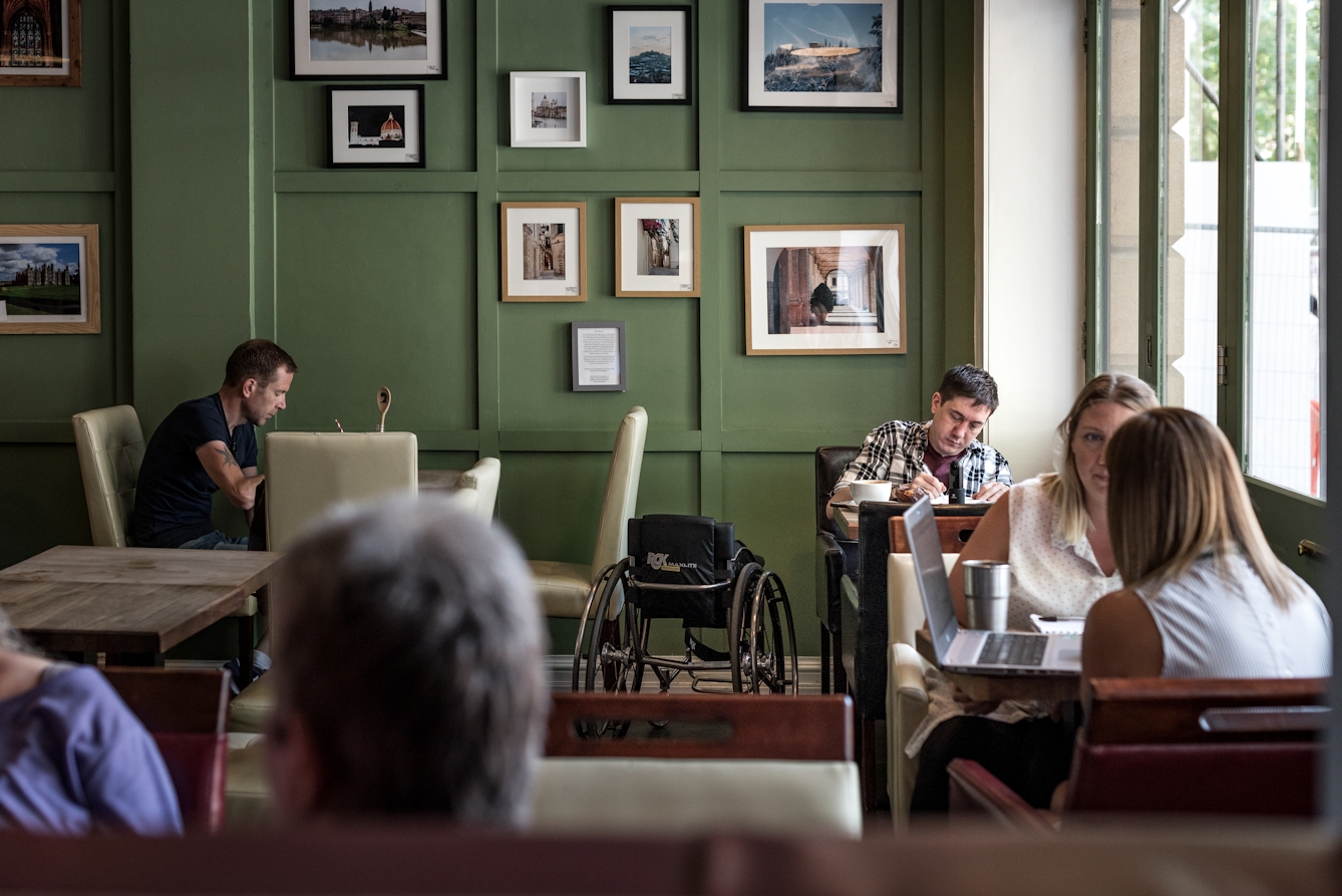 Photograph of a man with a coffee in a cafe working at a table. next to him is his wheelchair.