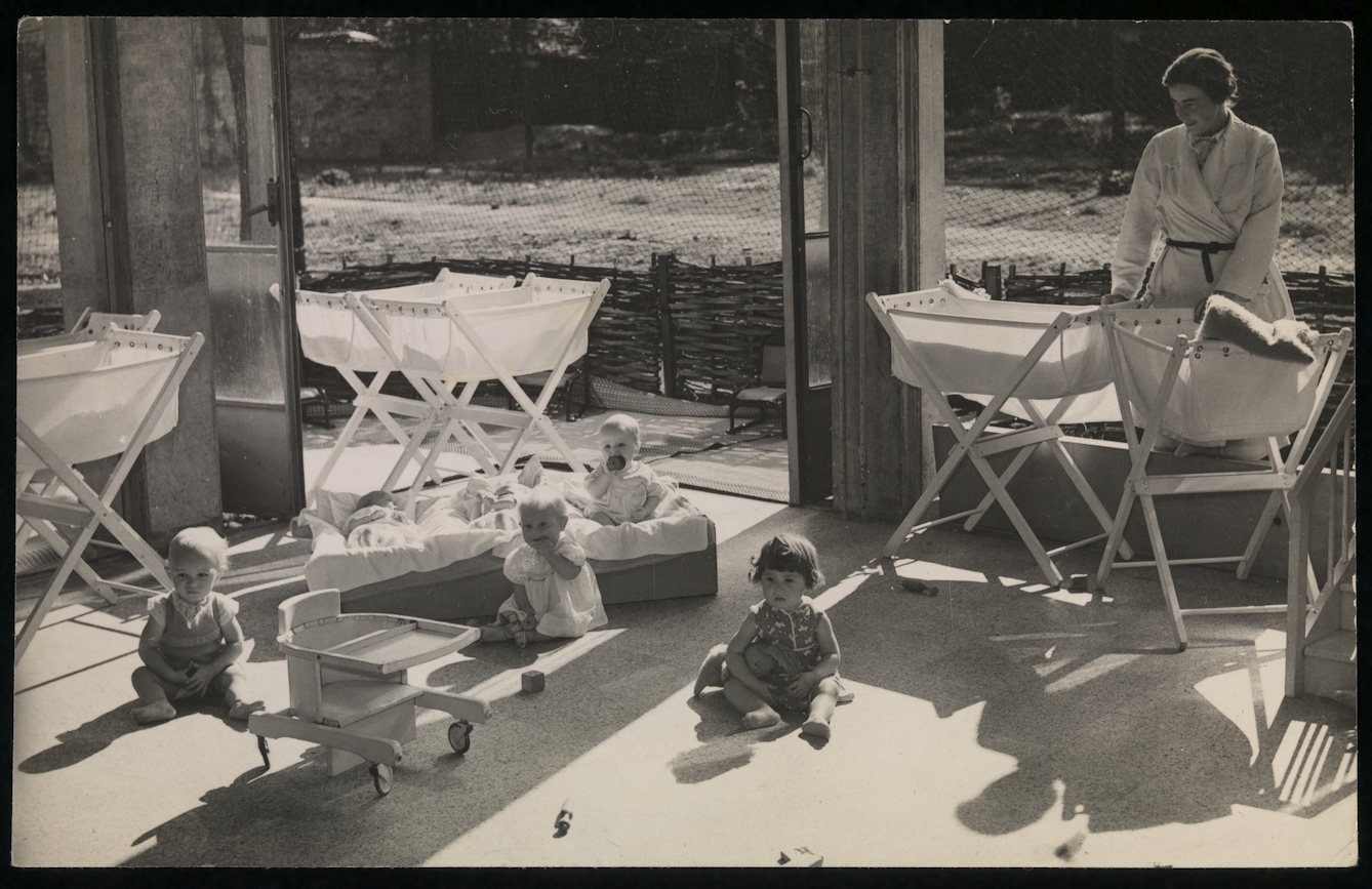 Black and white photograph showing a nursery, with several small children and a female nurse.