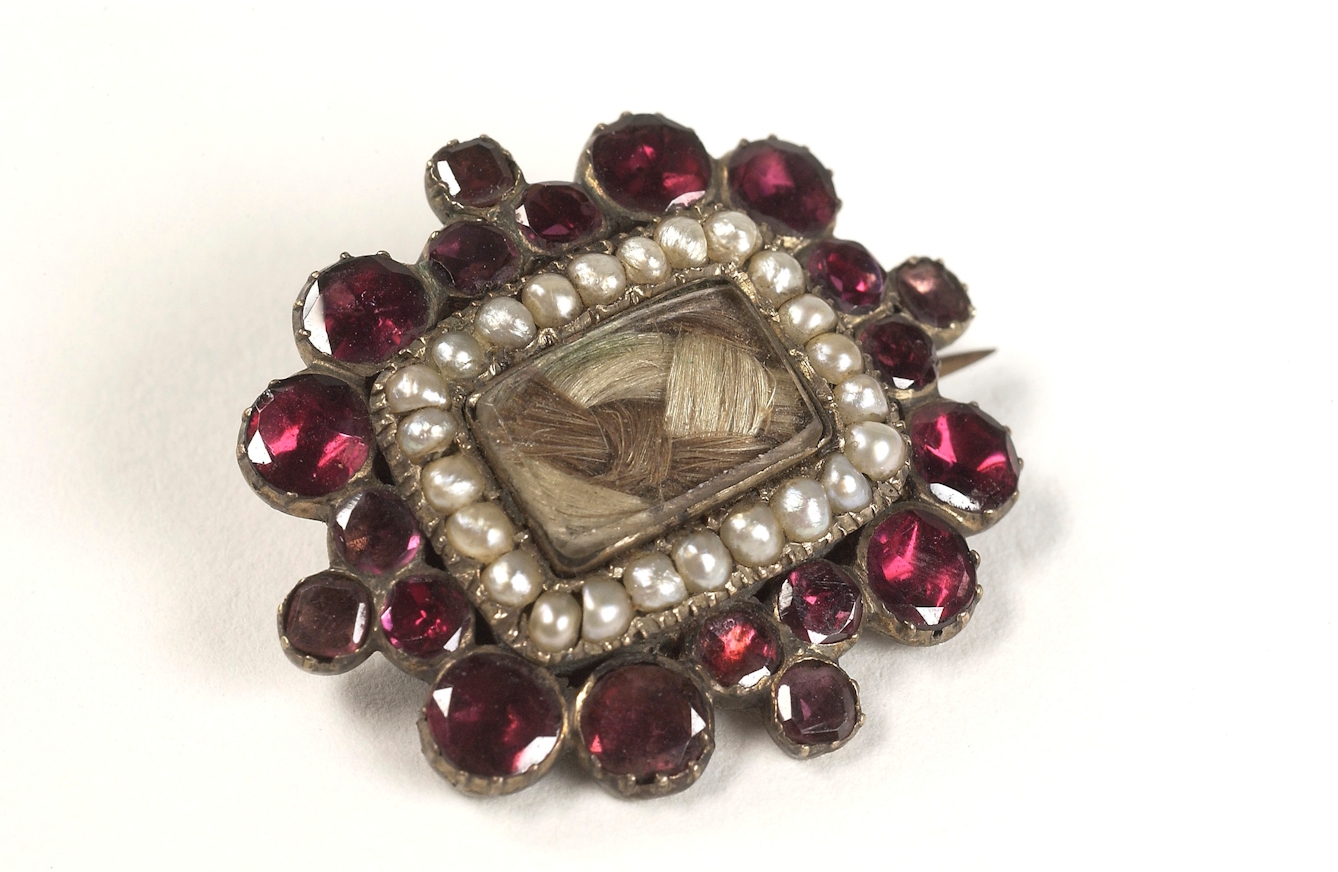 A rectangular brooch with human hair in the centre, surrounded by pearls and red gem stones.