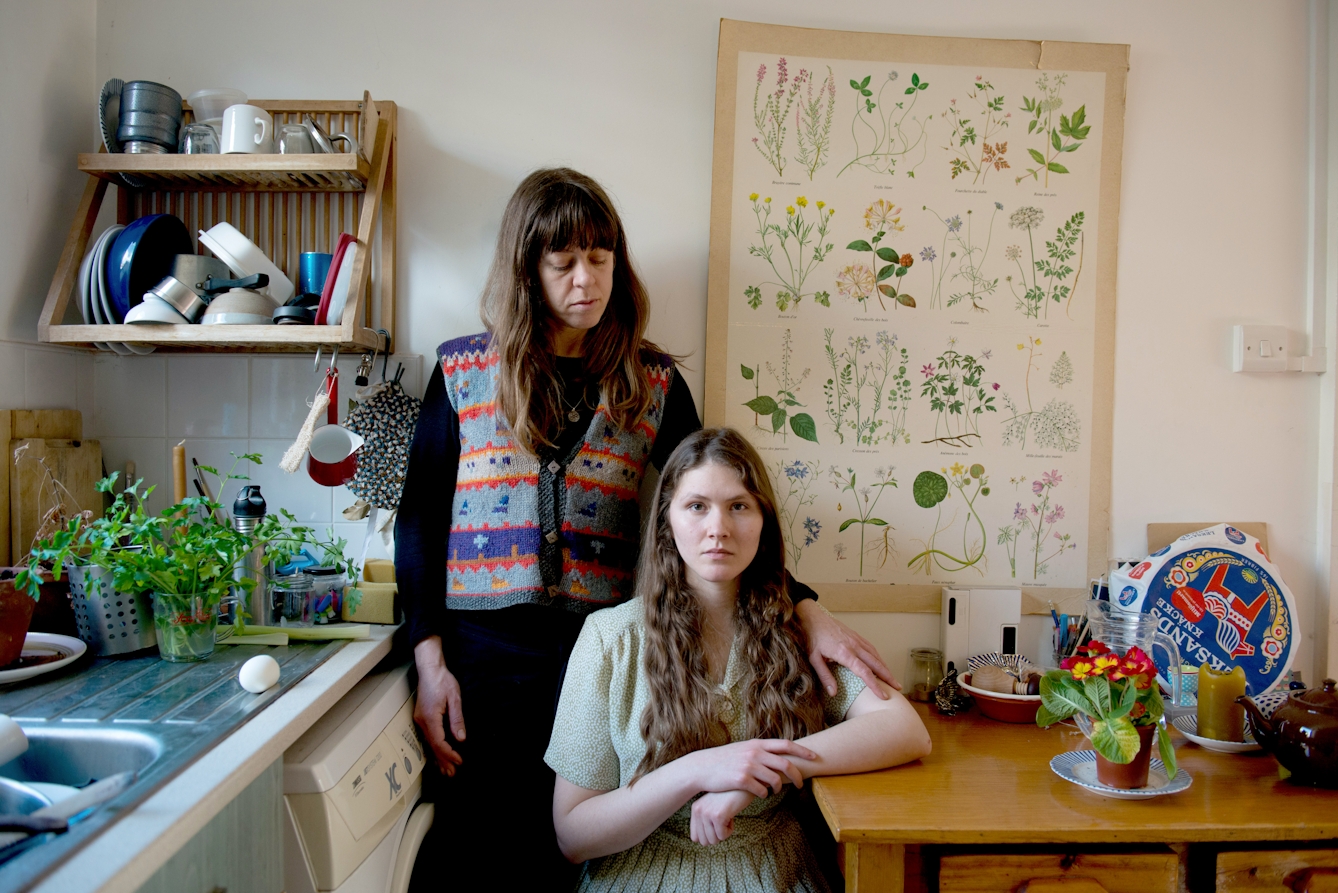 Photograph of a young woman and a mother figure in a domestic kitchen. The young woman is seated in a long dress and and the mother figure stands behind her, hand on her shoulder. The young woman is looking to camera, the mother figure is looking at her.